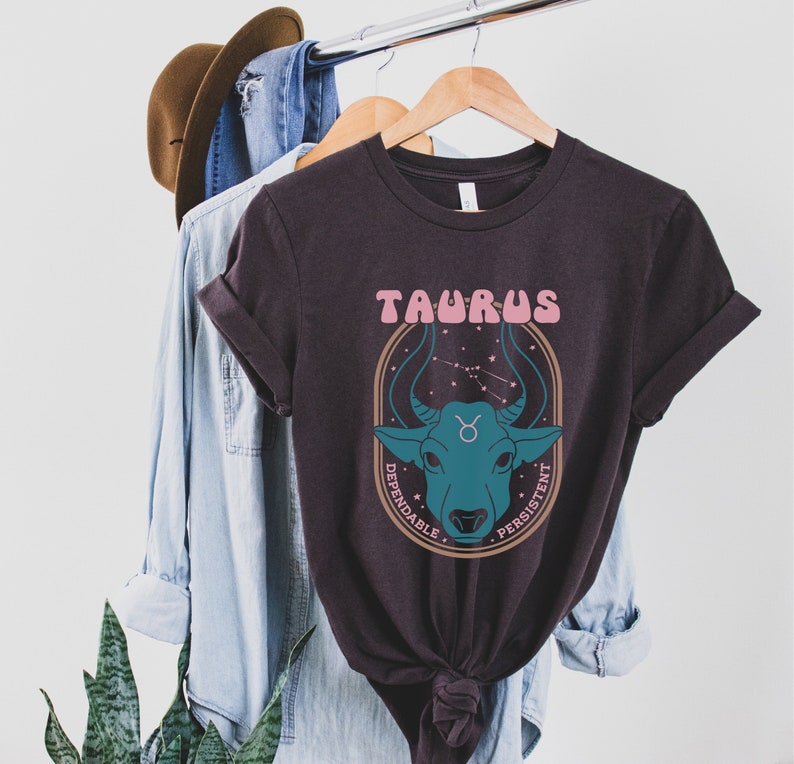K&ouml;nnte beinhalten: Ein dunkelbraunes T-Shirt mit einer Grafik des Tierkreiszeichens Stier. Das Zeichen ist ein Stierkopf mit einem rosa-blauen Farbschema. Der Text "Stier" ist in rosa Buchstaben &uuml;ber dem Stierkopf geschrieben. Die W&ouml;rter "Zuverl&auml;ssig" und "Beharrlich" sind in einem Kreis um den Stierkopf geschrieben.