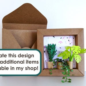 May include: A brown paper frame with a white background and a pink and black terrazzo pattern. Inside the frame are three paper plants in pots. The plants are green and have a 3D effect. The frame is sitting on a white surface. Create this design using additional items available in my shop!