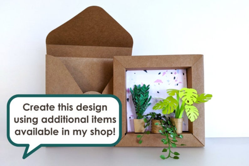 May include: A brown paper frame with a white background and a pink and black terrazzo pattern. Inside the frame are three paper plants in pots. The plants are green and have a 3D effect. The frame is sitting on a white surface. Create this design using additional items available in my shop!