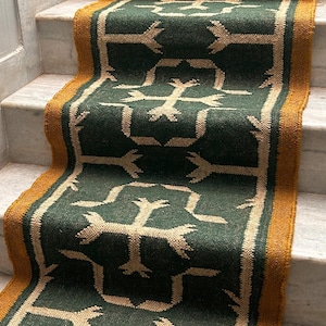 May include: A green and tan patterned stair runner with a gold border. The runner features a repeating geometric design. The runner is installed on white marble stairs. The text "CARPETEMPIRE" is visible on the bottom right corner.