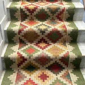 以下が含まれることがあります： 赤、緑、黄色、ベージュの色合いの幾何学的なひし形模様の階段ランナーラグ。ラグは緑色の縁取りがあり、白い大理石の階段に設置されています。「CARPETEMPIRE」の文字が下部に表示されています。