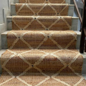 Puede incluir: Alfombra de escalera hecha de fibra de yute natural con un patrón de diamantes en beige y blanco roto. La alfombra está instalada en escaleras de mármol. El texto "CARPETEMPIRE" es visible en la parte inferior de la imagen.