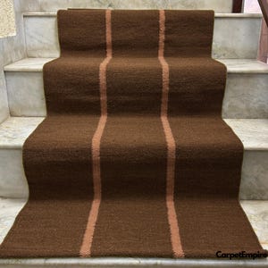 May include: A brown stair runner with three vertical stripes in a lighter shade of brown. The runner is made of a woven material and is placed on marble stairs. The runner adds a decorative element to the staircase.