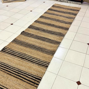 Op de afbeelding: Een vloerkleed van natuurlijke vezels met zwarte en bruine strepen. Het vloerkleed is gemaakt van jute en is perfect om elke kamer een vleugje natuurlijke textuur te geven.