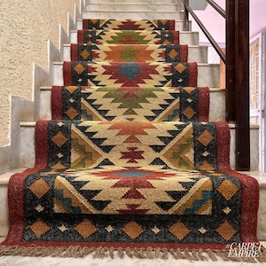 Può includere: Un set di pedate per scale con motivi geometrici colorati in tonalità di rosso, marrone, beige e blu. Le pedate per scale sono realizzate in juta tessuta e hanno un bordo a frangia. #CARPETEMPIRE