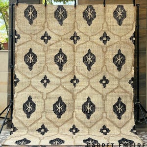 Puede incluir: Una alfombra beige rectangular grande con un patrón geométrico repetido en negro y beige. El diseño presenta formas de diamante y cruz. Las palabras "CARPET EMPIRE" están impresas en la parte inferior de la alfombra.