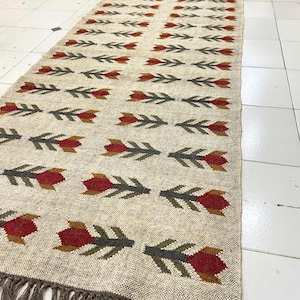 Puede incluir: Una alfombra larga y rectangular con fondo crema y un patrón repetitivo de flores estilizadas en tonos rojos, naranjas y verdes. La alfombra tiene un borde de flecos.