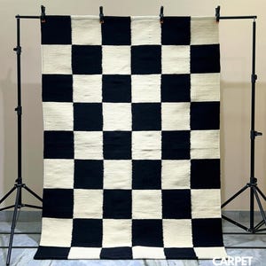 Puede incluir: Una alfombra rectangular grande con un llamativo diseño de tablero de ajedrez en negro y blanco roto. La alfombra se muestra verticalmente, suspendida de un marco de metal negro. La textura parece tejida y el diseño es simétrico.