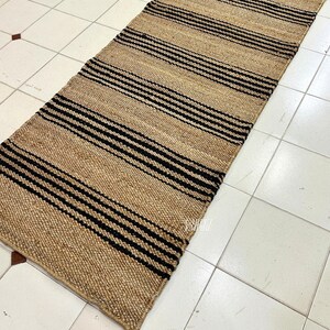Op de afbeelding: Een vloerkleed van natuurlijke vezels met zwarte en bruine strepen. Het vloerkleed is gemaakt van jute en is perfect om elke kamer een vleugje natuurlijke textuur te geven.