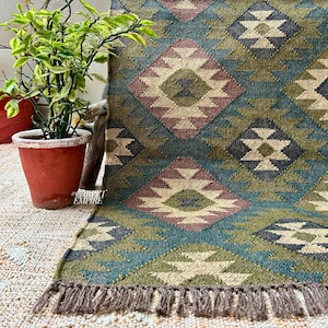 Puede incluir: Una alfombra geométrica con estampado verde, azul y marrón con flecos. La alfombra está hecha de tejido tejido y tiene un diseño tribal.