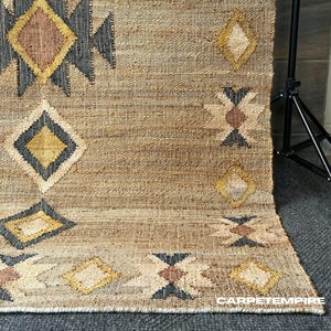 Pode incluir: Um tapete de juta bege com um padrão geométrico. O design apresenta formas de diamante e seta em preto, amarelo e castanho. O tapete tem uma textura tecida e uma estética natural e terrosa.