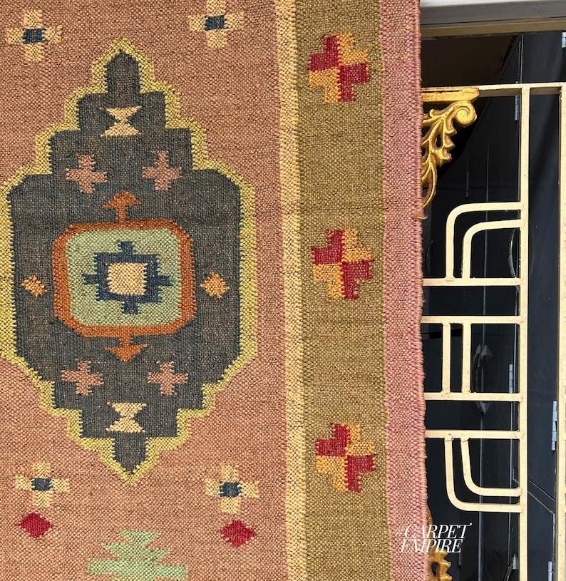 May include: A pink and brown rug with a geometric pattern in shades of blue, green, yellow, and orange. The rug has a fringe border and is made of wool.