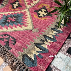 May include: A close-up of a vibrant, patterned rug with a rich magenta base. The rug features geometric designs in teal, black, beige, and yellow. The rug has a fringed edge and is likely made of woven material.