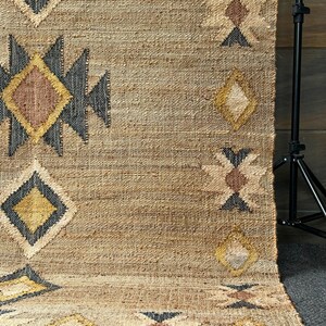 Natural fiber jute rug with beige base, cream geometric motifs, and a purplish‑black border framing design; handwoven with tight, flatweave technique. Brings texture and artisan feel to spaces like bedroom floor, living room or office area. Muted color helps to hide dust, the durable jute makes for long lasting use in moderate traffic areas.
