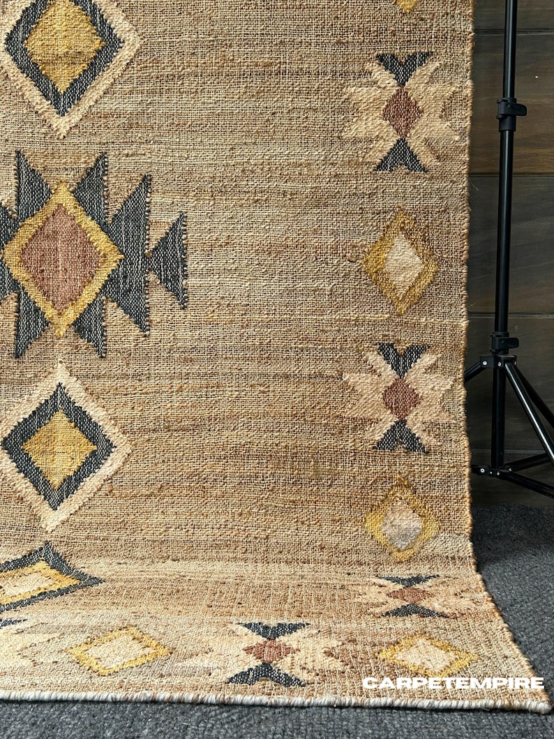 Natural fiber jute rug with beige base, cream geometric motifs, and a purplish‑black border framing design; handwoven with tight, flatweave technique. Brings texture and artisan feel to spaces like bedroom floor, living room or office area. Muted color helps to hide dust, the durable jute makes for long lasting use in moderate traffic areas.
