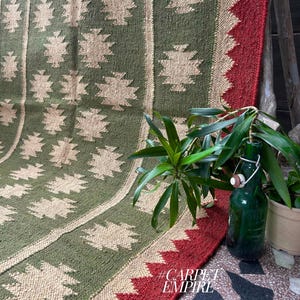 May include: A green and beige patterned rug with a red border. The rug features a repeating geometric design and is displayed with a green bottle and potted plants. The text "#CARPET EMPIRE" is visible in the lower right corner.