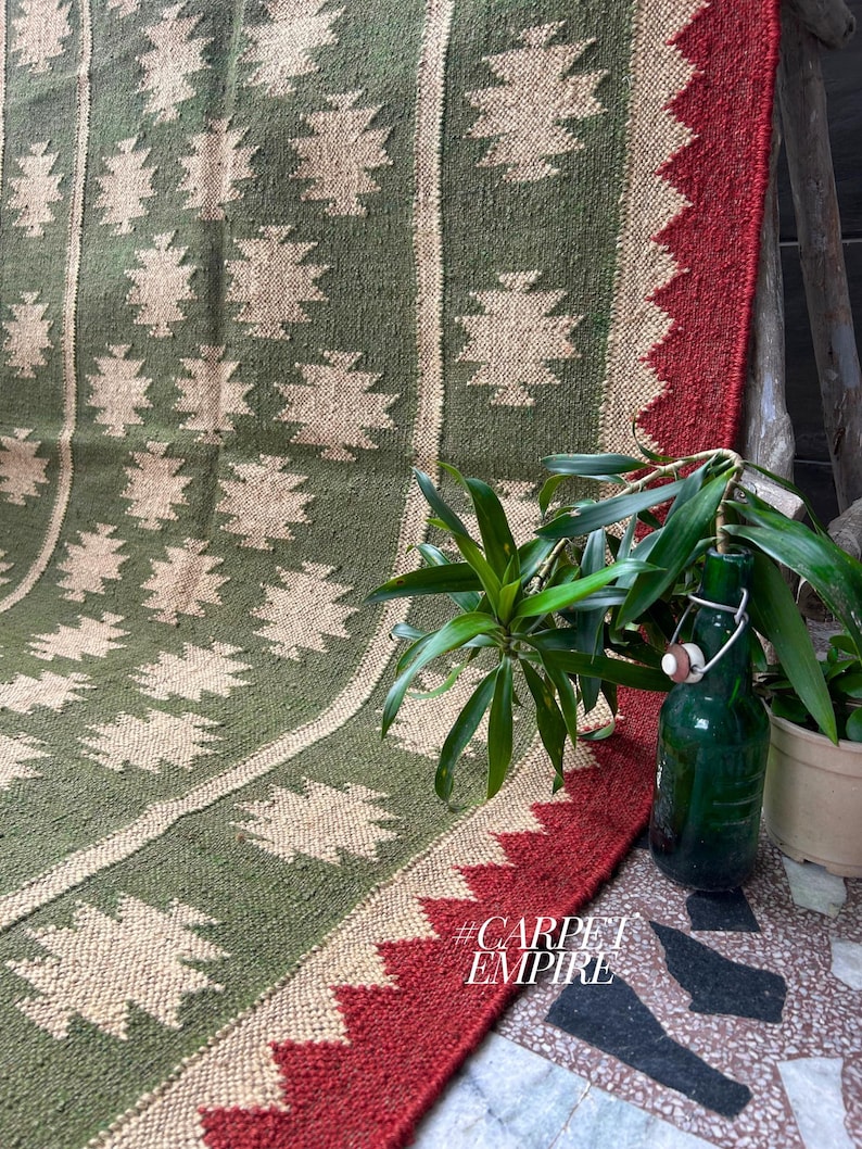 May include: A green and beige patterned rug with a red border. The rug features a repeating geometric design and is displayed with a green bottle and potted plants. The text "#CARPET EMPIRE" is visible in the lower right corner.