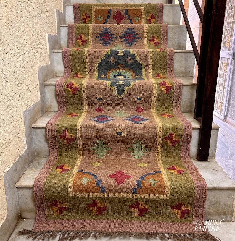 May include: A brown, green, and red patterned rug with a geometric design. The rug is laid out on a set of stairs. The rug has a fringe border. #CARPETEMPIRE