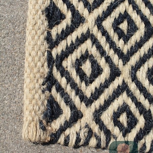 May include: Close-up of a black and natural jute rug with a geometric pattern. The rug has a woven texture and a frayed edge.