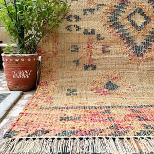 May include: A handwoven rug with a geometric pattern in shades of beige, pink, and blue. The rug has a fringe border and is made of natural fibres.
