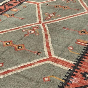 Sage green tribal kilim area rug with stacked terracotta outlined diamond medallions and scattered cross motifs. Ornate border with fringe. Wool jute flatweave. Custom.