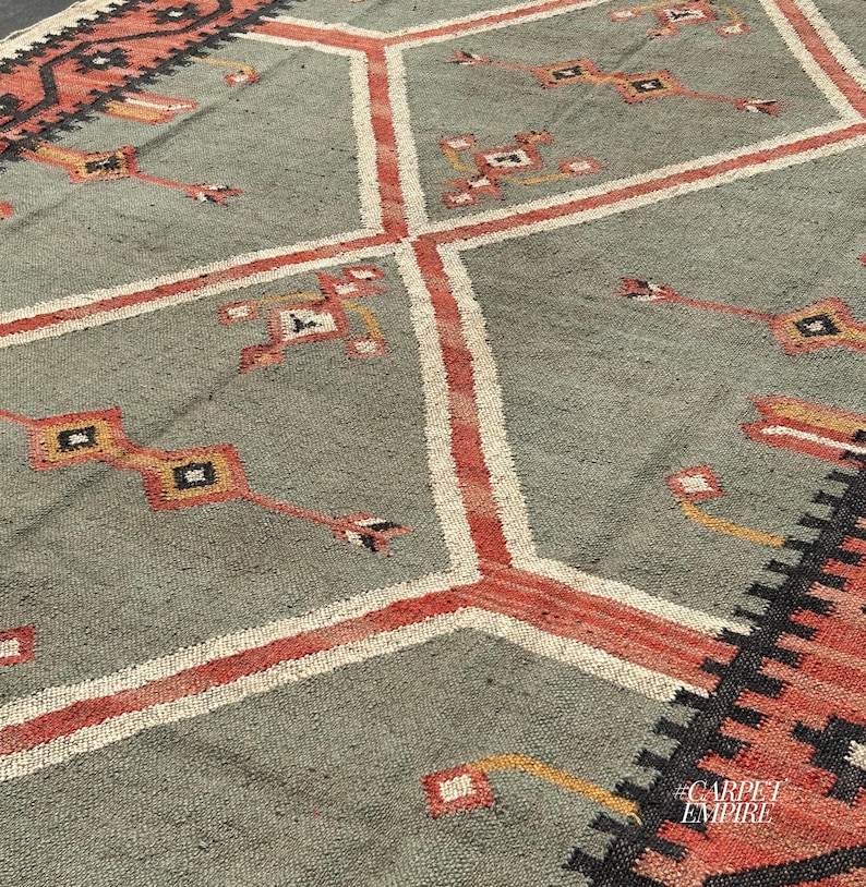 Sage green tribal kilim area rug with stacked terracotta outlined diamond medallions and scattered cross motifs. Ornate border with fringe. Wool jute flatweave. Custom.