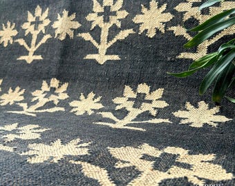 May include: Close-up of a dark blue rug featuring a repeating floral pattern in a light beige hue. The rug has a textured appearance and is made of a woven material. The floral design is stylized and symmetrical, creating a decorative element.