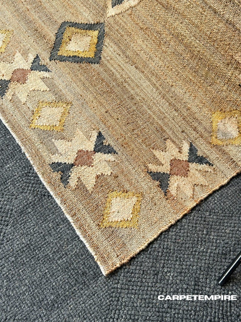 Handwoven natural jute area rug with a beige base, centered cream kilim designs, and a gentle purplish‑black border edge. The flatweave structure ensures low profile and easy cleaning; durable fibre makes it a smart choice for moderate traffic in living spaces or bedrooms. Muted patterning adds warmth and interest without overpowering other design elements.