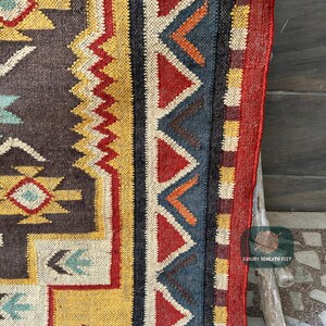 May include: Close-up of a colorful woven rug with a geometric pattern in shades of red, yellow, blue, and brown. The rug has a fringe border and is made of natural fibers.