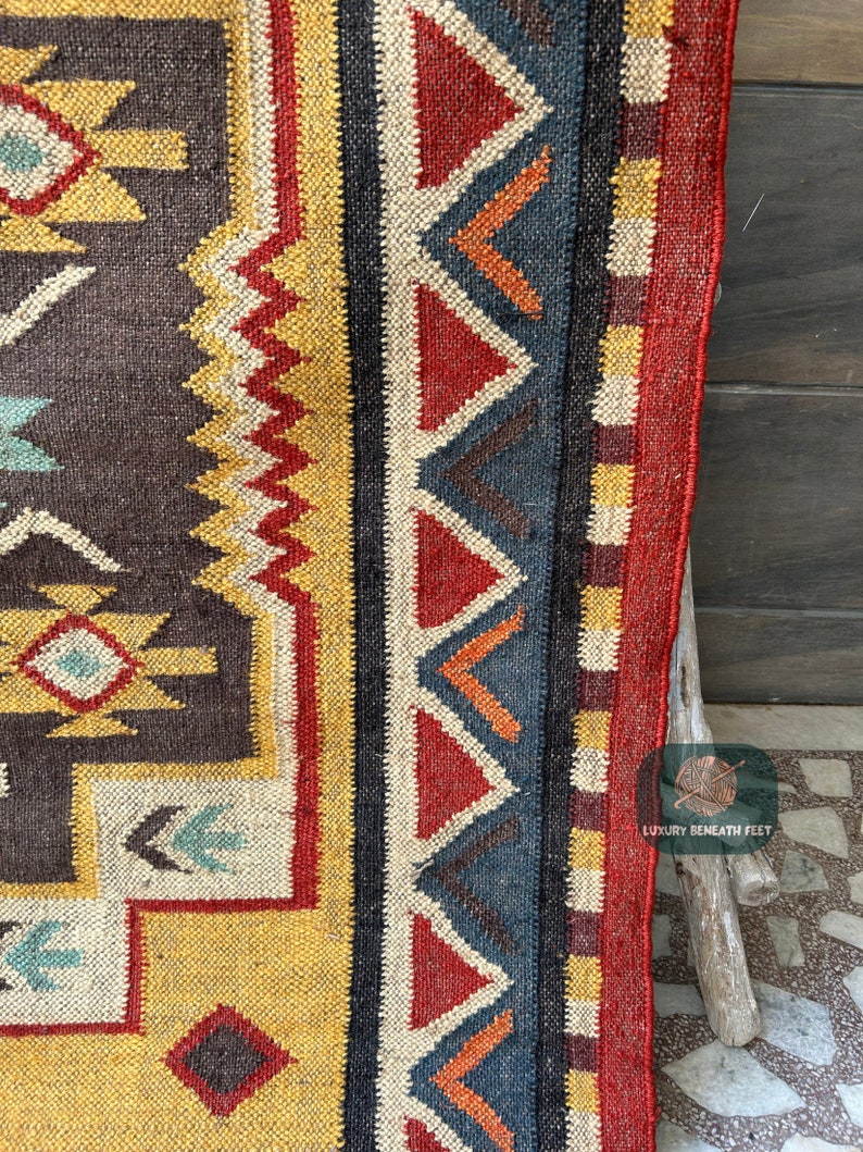 May include: Close-up of a colorful woven rug with a geometric pattern in shades of red, yellow, blue, and brown. The rug has a fringe border and is made of natural fibers.