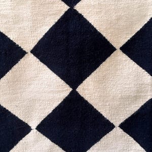 Modern geometric wool rug with black and white diamond check pattern. The handcrafted flatweave texture gives a clean and minimalist look, ideal for Scandinavian or neutral home decor. Made from 100% wool for comfort and durability, this rug complements living rooms, bedrooms, and minimalist office spaces.