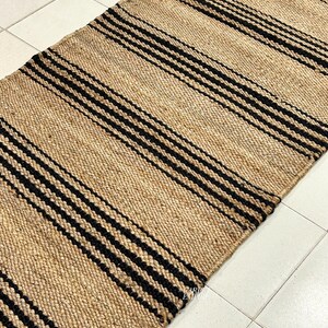 Op de afbeelding: Een vloerkleed van natuurlijke vezels met zwarte strepen. Het vloerkleed is gemaakt van jute en heeft een geweven textuur.