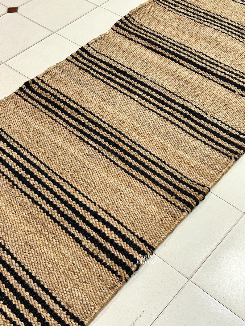 Op de afbeelding: Een vloerkleed van natuurlijke vezels met zwarte strepen. Het vloerkleed is gemaakt van jute en heeft een geweven textuur.