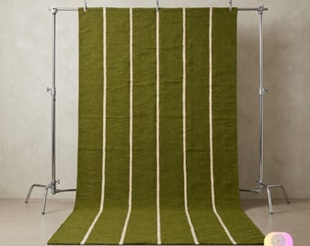 May include: A rectangular area rug featuring a solid olive green base with vertical cream-colored stripes running from top to bottom. The rug is suspended from a metal stand with clips along the top edge, displaying a flat, woven texture.
