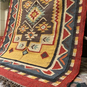 May include: A hand-woven rug with a geometric pattern in shades of red, yellow, blue, and brown. The rug has a fringe border and is made of natural fibers.