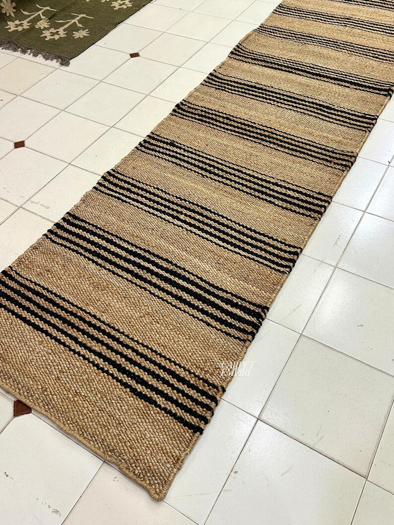 Op de afbeelding: Een vloerkleed van natuurlijke vezels met zwarte en bruine strepen. Het vloerkleed is gemaakt van jute en is perfect om elke kamer een vleugje natuurlijke textuur te geven.