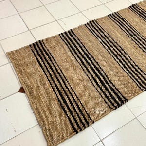 Op de afbeelding: Een vloerkleed van natuurlijke vezels met zwarte en bruine strepen. Het vloerkleed is geweven met een eenvoudig patroon en heeft een natuurlijke, rustieke uitstraling.