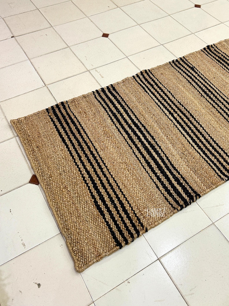 Op de afbeelding: Een vloerkleed van natuurlijke vezels met zwarte en bruine strepen. Het vloerkleed is geweven met een eenvoudig patroon en heeft een natuurlijke, rustieke uitstraling.