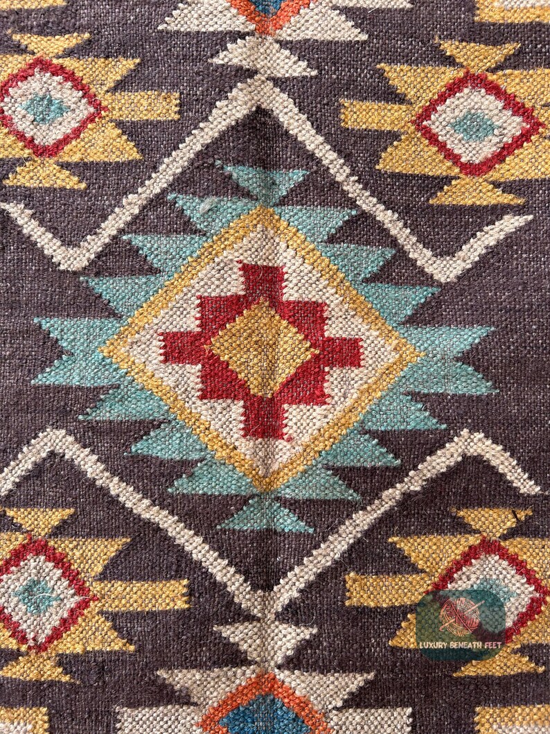May include: Close-up of a brown woven rug with a geometric pattern in shades of red, yellow, blue, and white. The pattern features triangles, diamonds, and squares.