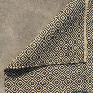May include: A black and natural jute rug with a geometric diamond pattern. The rug is folded in half, showing the pattern on both sides. The rug is on a grey concrete surface. The text "Luxury Beneath Feet" is visible in the bottom right corner of the image.