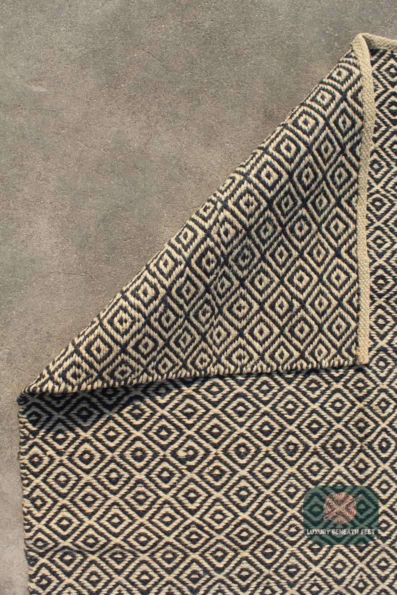 May include: A black and natural jute rug with a geometric diamond pattern. The rug is folded in half, showing the pattern on both sides. The rug is on a grey concrete surface. The text "Luxury Beneath Feet" is visible in the bottom right corner of the image.
