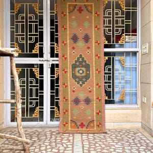 May include: A long, narrow rug with a pink background and intricate geometric patterns in shades of red, blue, green, and yellow. The rug is hanging on a wall in front of a door with a decorative iron grate.