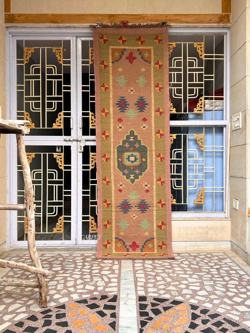 May include: A long, narrow rug with a pink background and intricate geometric patterns in shades of red, blue, green, and yellow. The rug is hanging on a wall in front of a door with a decorative iron grate.