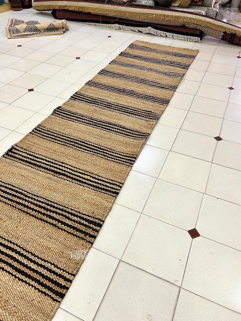 Op de afbeelding: Een vloerkleed van natuurlijke vezels met zwarte en bruine strepen. Het vloerkleed is gemaakt van jute en is perfect om elke kamer een vleugje natuurlijke textuur te geven.