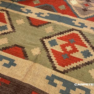 Op de afbeelding: Close-up van een geweven tapijt met geometrische patronen. Het tapijt heeft een kleurenpalet van rood, blauw, beige en groen. Het ontwerp omvat ruitvormen en lineaire elementen, waardoor een getextureerd oppervlak ontstaat.