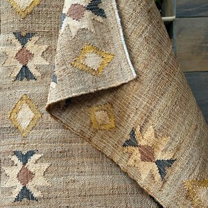 Natural jute rug showcasing cream geometric kilim motifs on a beige field with a subtle purplish‑black trimmed border. The flatweave jute fiber adds texture and tactile interest, while the muted colors blend effortlessly into neutral decor schemes. This area rug delivers visual structure with its pattern without overwhelming softness or brightness—suited for living rooms, hallways, or spaces that emphasize calm, grounded design.