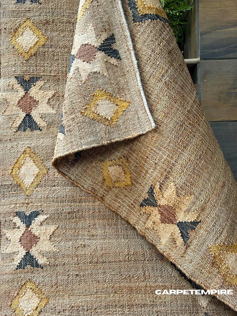 Natural jute rug showcasing cream geometric kilim motifs on a beige field with a subtle purplish‑black trimmed border. The flatweave jute fiber adds texture and tactile interest, while the muted colors blend effortlessly into neutral decor schemes. This area rug delivers visual structure with its pattern without overwhelming softness or brightness—suited for living rooms, hallways, or spaces that emphasize calm, grounded design.
