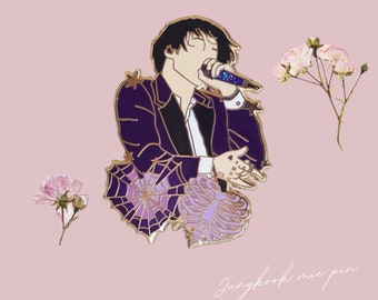 May include: A gold-tone enamel pin featuring a purple-clad figure holding a microphone, surrounded by purple and gold spiderwebs and flowers. The text "Jungkook mic pin" is written below the figure.