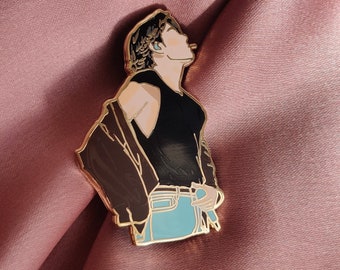 Puede incluir: Pin de esmalte dorado con una ilustración de dibujos animados de una persona que lleva un top negro sin mangas, una chaqueta marrón y unos vaqueros azules. La persona mira hacia la derecha y tiene el pelo negro.