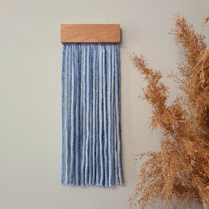 May include: A blue and white yarn wall hanging with a wooden top. The yarn is woven into a fringe that hangs down from the wooden piece.